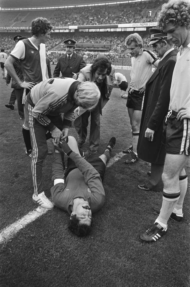 Schwarz-weißes Bild eines Fußballspielers, der auf dem Boden liegt und von einer Gruppe Männer umgeben ist, mit einer Zuschauermenge im Hintergrund eines Stadions.