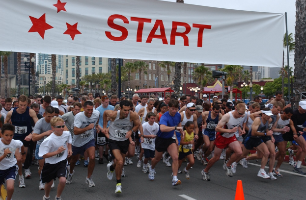 Gruppe von Menschen, die bei einem Marathon laufen, mit einem Verkehrskegel im Vordergrund und einem Banner im Hintergrund, umgeben von Bäumen, Laternenmasten, Gebäuden und einem klaren blauen Himmel.