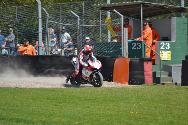 Eine Person in Helm und Handschuhen fährt ein Motorrad über ein grasbewachsenes Feld mit Zuschauern, Absperrungen, Treppen und einem nummerierten Schuppen im Hintergrund.