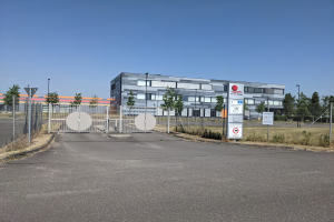 Ein großes Gebäude mit vielen Fenstern, beschriftet mit First Solar, umgeben von einem Metallzaun, Bäumen, Pflanzen, Gras, Schildern, Straßenmasten und einem Weg.