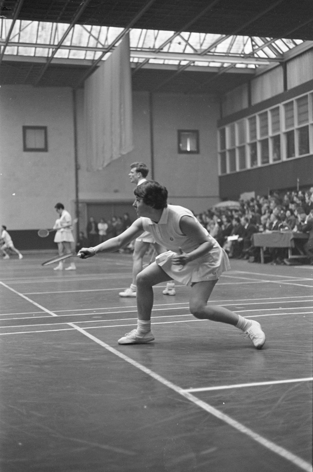 Schwarzes und weißes Foto von zwei Frauen, die Badminton auf einem Court spielen, jede hält einen Schläger in der Hand, mit Zuschauern rechts und einer fensterumrahmten Wand im Hintergrund.