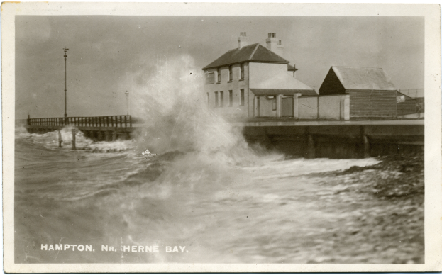 Eine große Welle kracht gegen eine Mole auf einem Schwarz-Weiß-Foto, mit Häusern, Pfählen und einer Brücke im Hintergrund und Text am unteren Bildrand.