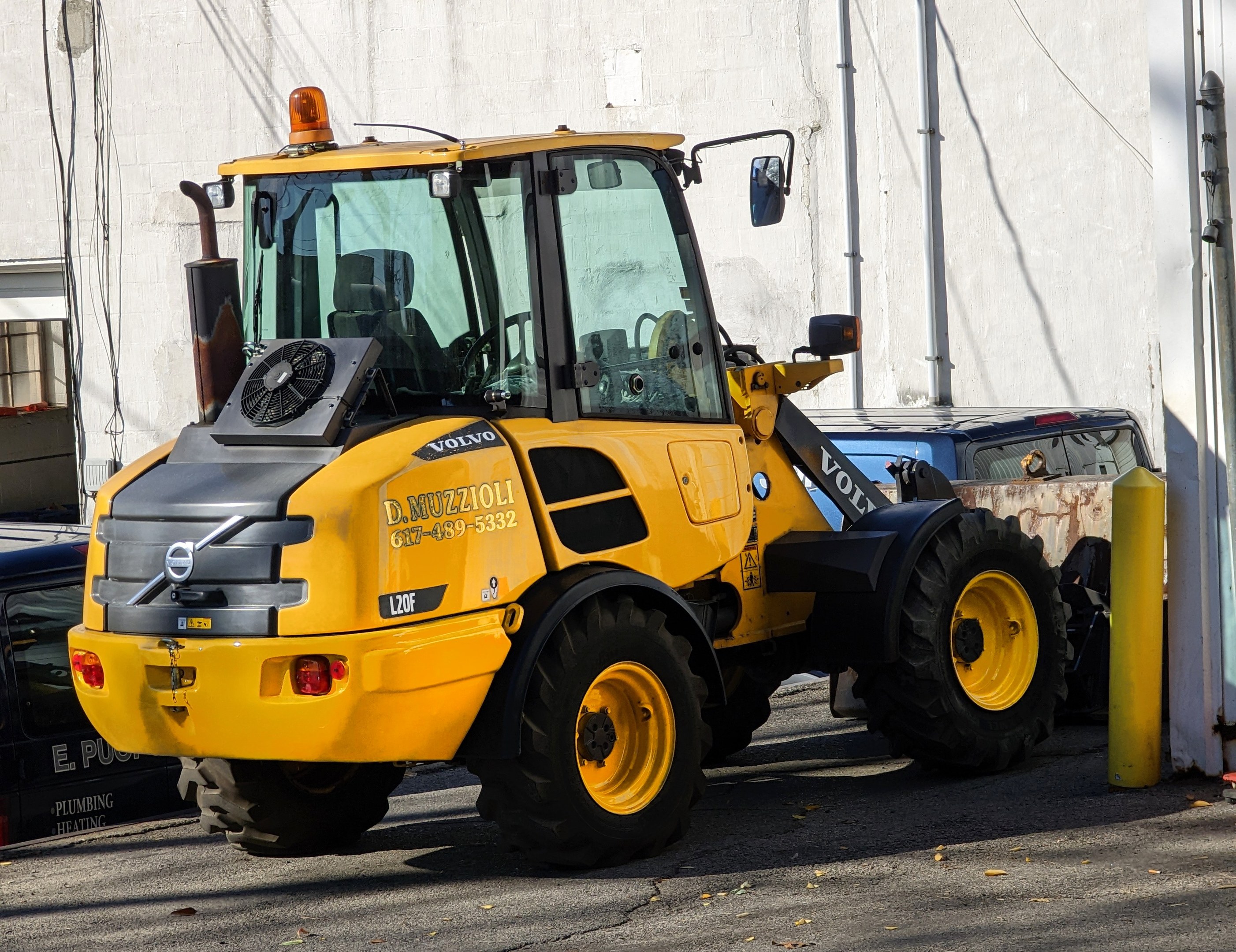 Ein gelber Radlader steht auf einer Straße vor einem Gebäude mit einer Wand im Hintergrund, mit anderen Fahrzeugen in der Nähe.
