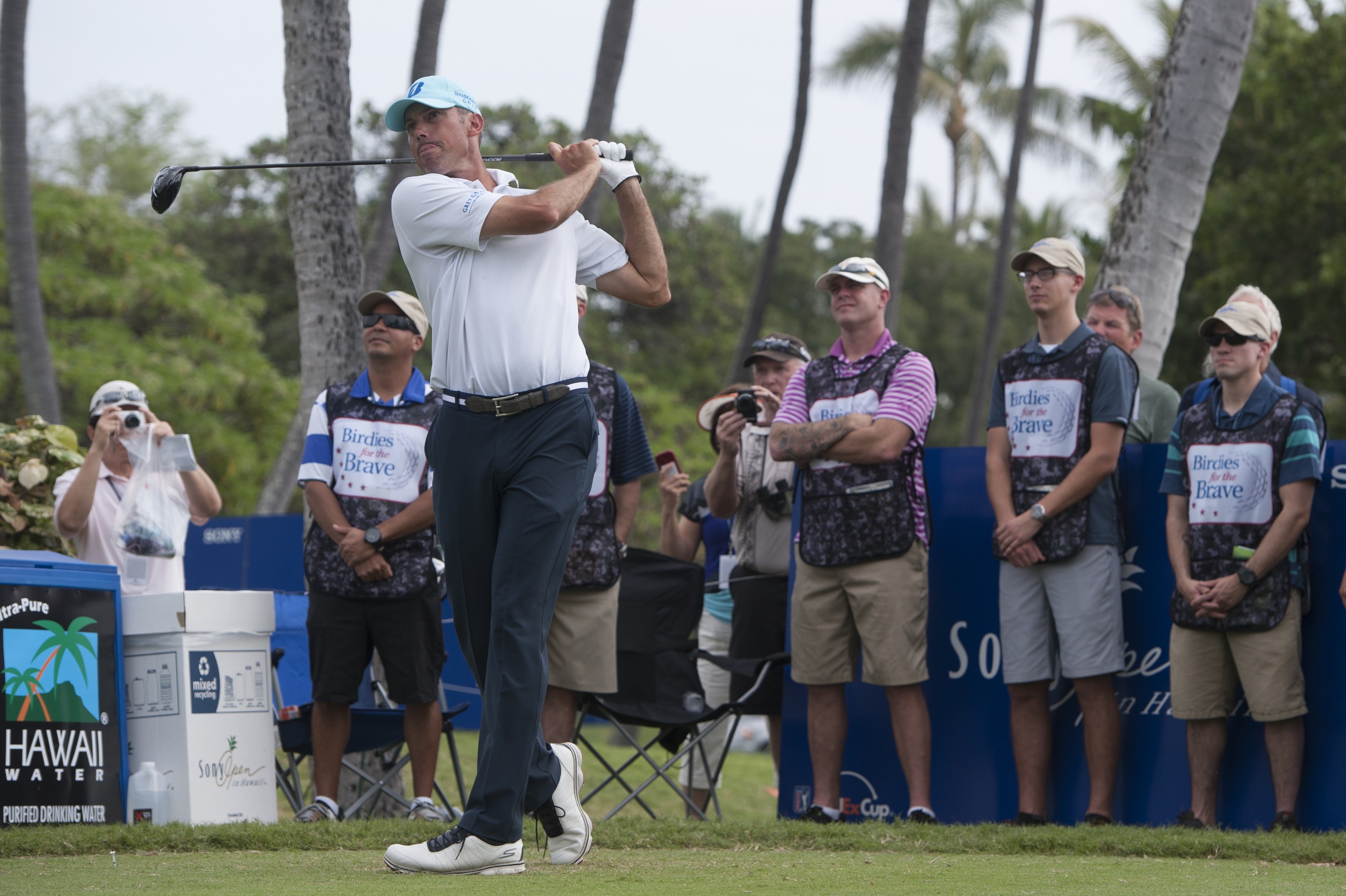 Ein Mann in einem weißen T-Shirt, schwarzen Hosen und einer blauen Mütze schwingt einen Golfschläger auf einem grünen Feld, während Zuschauer mit Kameras beobachten, mit Bäumen und einem klaren blauen Himmel im Hintergrund.