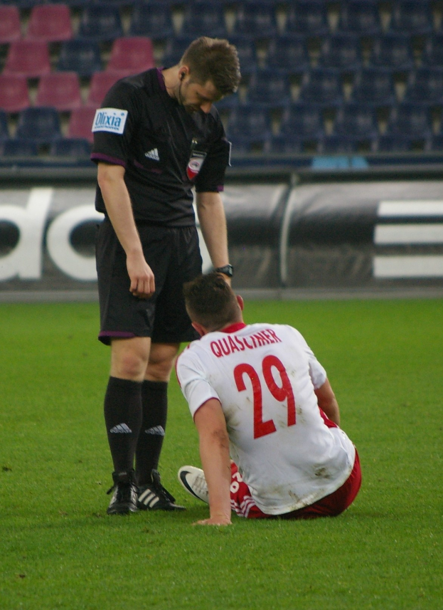 Ein Fussballspieler und ein Schiedsrichter sitzen auf dem Boden in einem Stadion, beide in Sportkleidung.