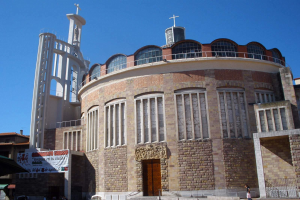 Die Kirche des Heiligen Grabes, ein großes Gebäude mit einem zentralen Uhrturm, umgeben von Fenstern, einer Tür, einer Fahne, einem Metallzaun, einer Gruppe von Menschen, Fahrzeugen, Zelten und einem bewölkten Himmel.