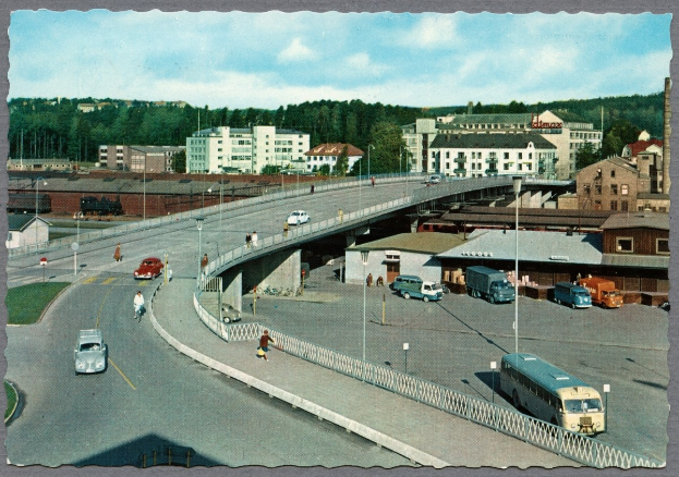 Altes Schwarz-Weiß-Foto einer Stadtstraße mit Fahrzeugen, Fußgängern auf einer Brücke, Laternen, Gebäuden, Bäumen und einem bewölkten Himmel.