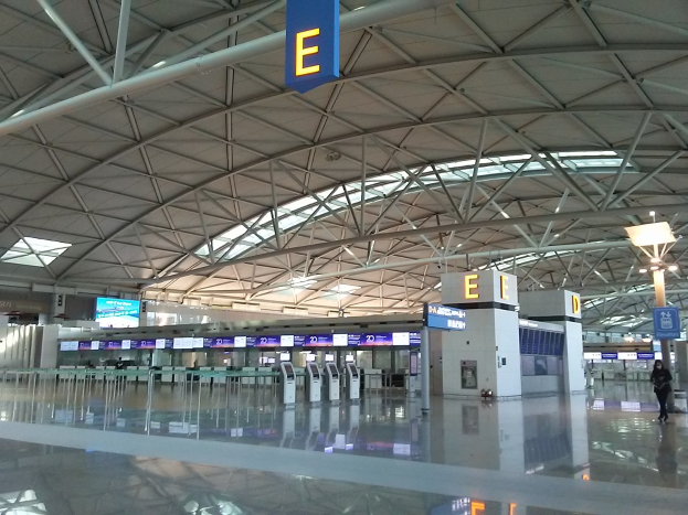 Leerer Flughafen-Terminal mit Menschen, die gehen, Schildern mit Text und Maschinen in der Mitte, einer Person links und Metallstangen und Lampen an der Decke.