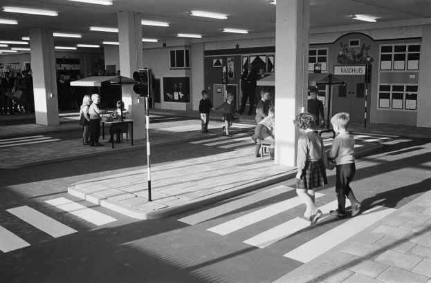 Schwarz-weißes Foto von Kindern, die eine Straße an einer Ampel überqueren, mit Menschen auf Bänken, einem Verkehrssignalmast im Vordergrund und Säulen mit Deckenlampen im Hintergrund.