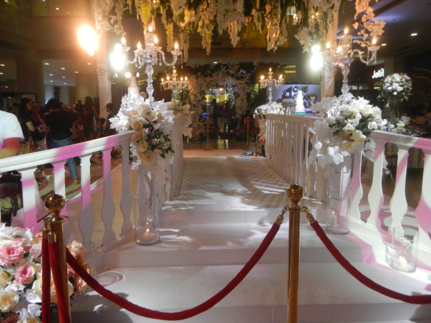 Eine Hochzeitszeremonie in einem Einkaufszentrum mit weißem Teppich, verzierten Geländern, Blumenarrangements und Deckenbeleuchtung, bei der zahlreiche Gäste anwesend sind.