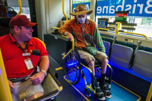 Zwei Männer im Rollstuhl auf einem Bus, einer trägt ein rotes T-Shirt und eine Kappe, der andere ein orangefarbenes Hemd und einen braunen Hut, beide tragen Masken und Ausweise, mit gelben Stangen und Glasfenstern, durch die man Bäume, Gebäude und den Himmel sieht.