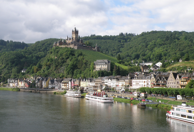 Ein malerischer Blick auf den Rhein in Deutschland mit einer Burg auf einem Hügel, Booten auf dem Fluss und Fahrzeugen auf einer näheren Straße unter einem bewölktem Himmel.