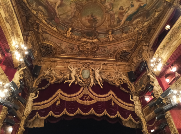 Theaterinnenraum mit roter Gardine, Kronleuchtern, Skulpturen, Lichtern, Deckenmalerei und einer Bühne mit einigen Menschen davor.