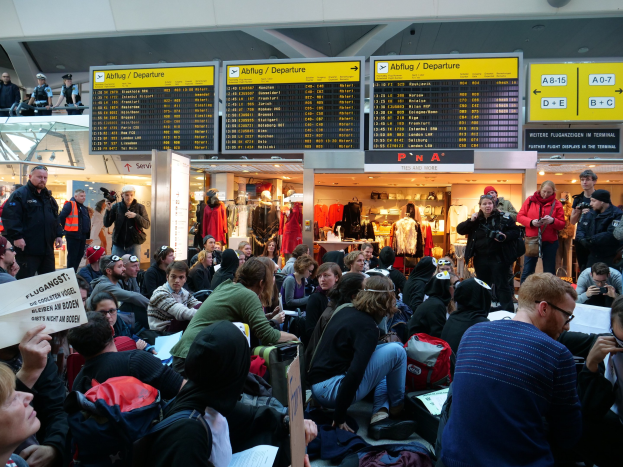 Eine große Gruppe von Menschen sitzt und steht in einem Flughafen während einer Protestaktion, mit Schildern, Puppen und Deckenleuchten im Hintergrund.