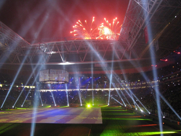 Olympiastadion in London bei Nacht, beleuchtet von hellen Lichtern und Feuerwerken, mit einem großen Bildschirm im Vordergrund und einer Menge drinnen.