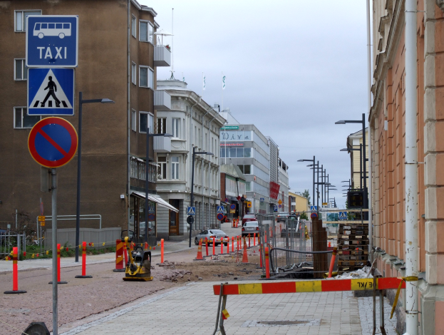 Eine Stadtstraße mit Gebäuden, Straßenlaternen, Straßenschildern, Verkehrszeichen, Verkehrskegeln, Kraftfahrzeugen, Absperrpollern, Bäumen und einem Himmel mit Wolken im Hintergrund.