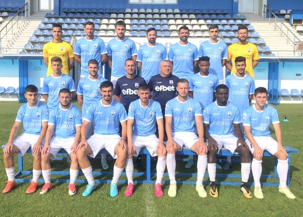 Männer in Sportkleidung stehend und sitzend auf einem Fußballfeld mit Tribünen und Gebäudestrukturen im Hintergrund.