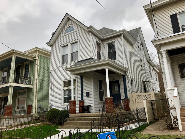 Reihe von Mietshäusern mit Fenstern, Türen, Säulen, Geländern, Treppen und Dächern, umgeben von einem Metallzaun, umgeben von Gras, Pflanzen, Bäumen und einem bewölkten Himmel, mit einem Mietschild am Boden, das die Adresse und die Listen Details anzeigt.