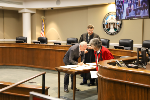 Eine Gruppe von drei Personen steht um einen Tisch in einem Gerichtssaal, unterschreibt Papiere, mit verschiedenen Gegenständen auf dem Tisch und Elementen wie Stühlen, einer Flagge, einem Logo, einem Fernseher und Deckenleuchten im Hintergrund.