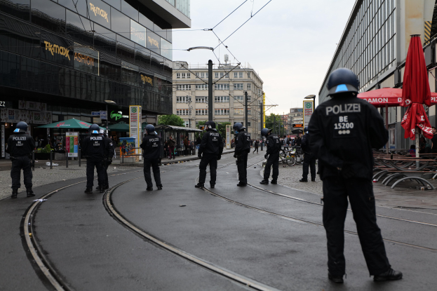Eine Gruppe von Polizeibeamten in schwarzen Uniformen und Helmen steht auf einer Straße, die von Glasfenster-Gebäuden gesäumt ist, mit Bäumen, Strommasten und verstreuten Outdoor-Möbeln im Hintergrund unter einem klaren Himmel.