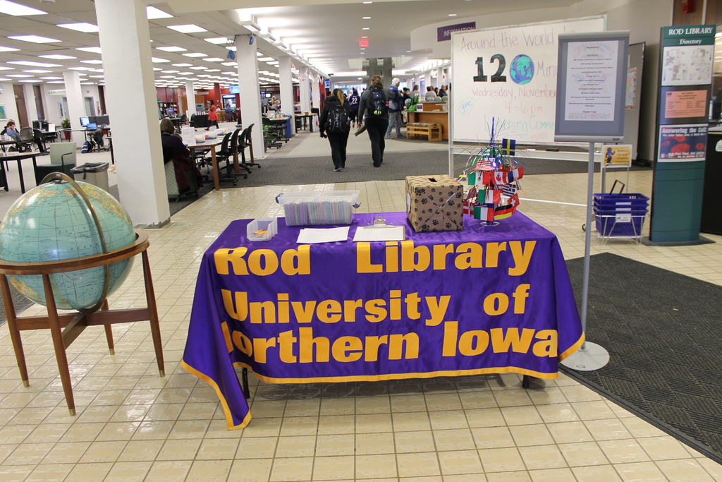 Eine Ausstellung mit einem Tisch, auf dem verschiedene Landesflaggen, eine Box, Papiere und eine Fahne sowie links daneben eine Weltkugel, rechts eine Anschlagtafel mit einigen Menschen in der Nähe und viele Tische und Stühle im Hintergrund zu sehen sind.