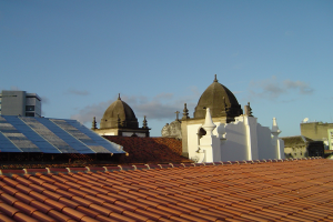 Stadtansicht mit mehreren Gebäuden im Vordergrund und einem klaren blauen Himmel im Hintergrund, die Solarpanels auf dem Dach eines Gebäudes zeigt.