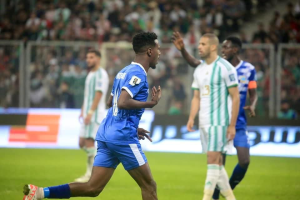 Ein Fussballspieler in einem blauen und weissen Trikot feiert mit erhobenen Armen vor einer Gruppe von Menschen in weissen und grünen Trikots, umgeben von einem leicht unscharfen Hintergrund mit einer weissen und blauen Tafel und einem Geländer.