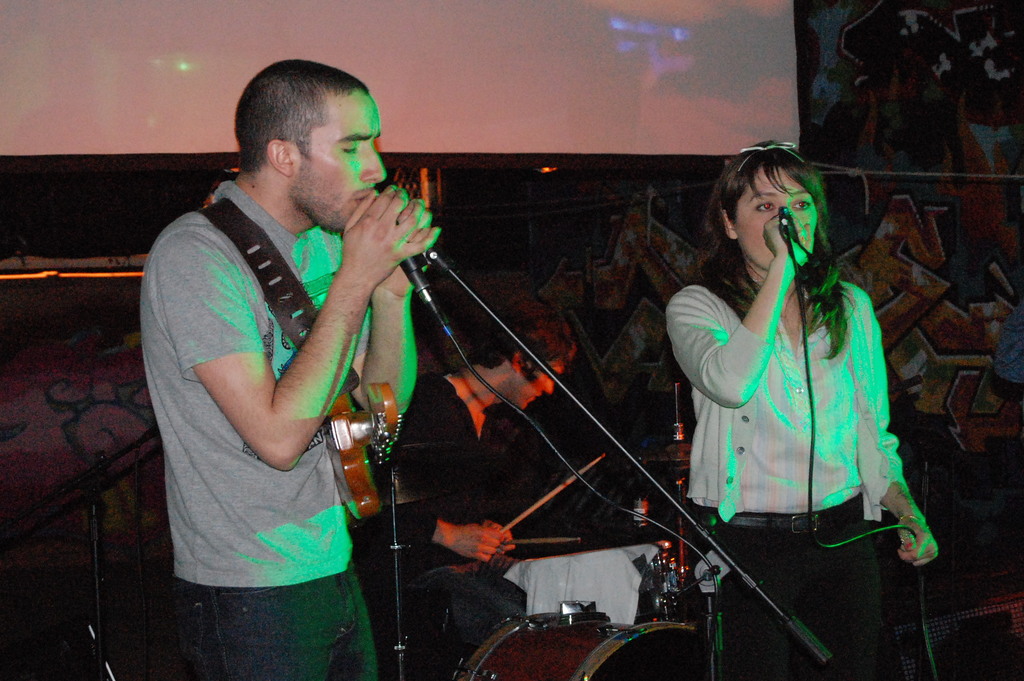 Ein Mann links und eine Frau rechts halten jeweils ein Mikrofon und singen, während eine Person in der Mitte ein Musikinstrument spielt.