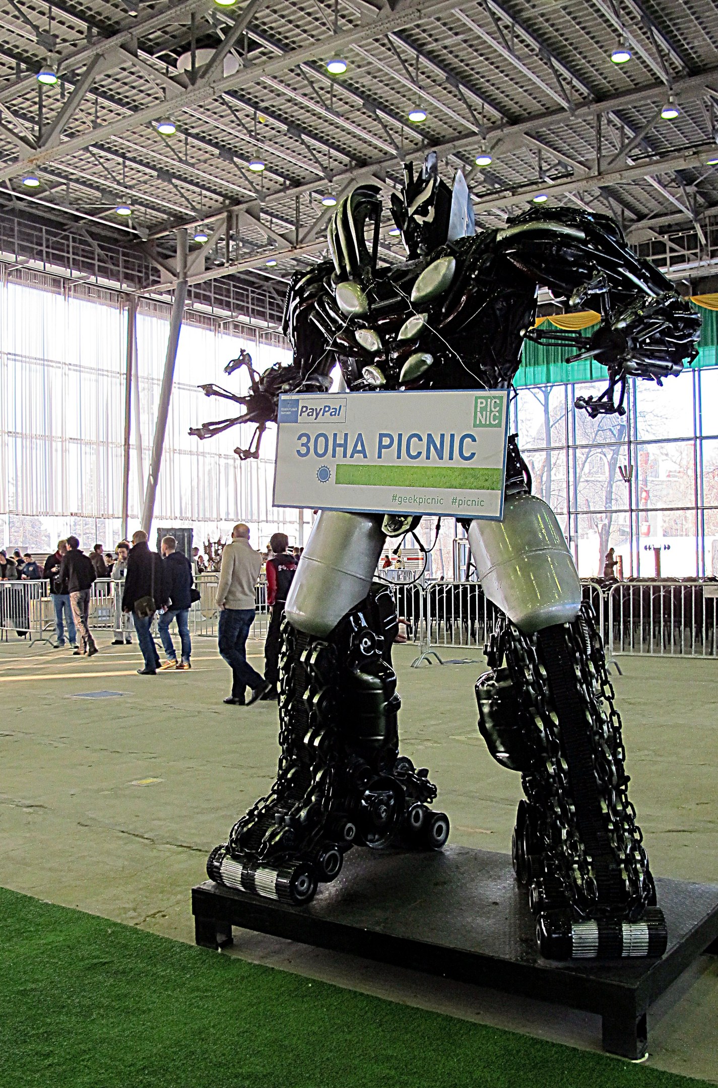 Große Metallroboterskulptur auf einem grünen Feld mit Menschen, einem Zaun, einem Schild, einem Dach mit Lampen und Metallpfählen.