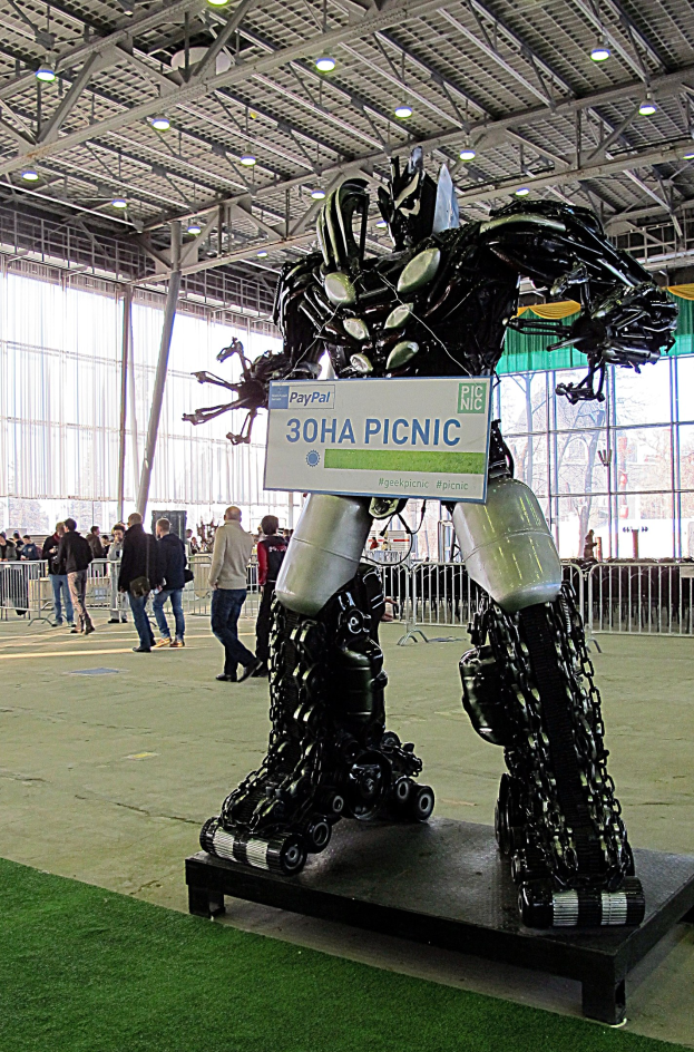 Große Metallroboterskulptur auf einem grünen Feld mit Menschen, einem Zaun, einem Schild, einem Dach mit Lampen und Metallpfählen.