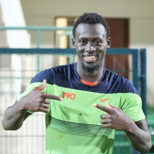 Ein Mann in einem grünen und blauen Hemd zeigt auf die Kamera und lächelt, steht in der Nähe einer Brüstung, einer Wand und einer Tür im Hintergrund.