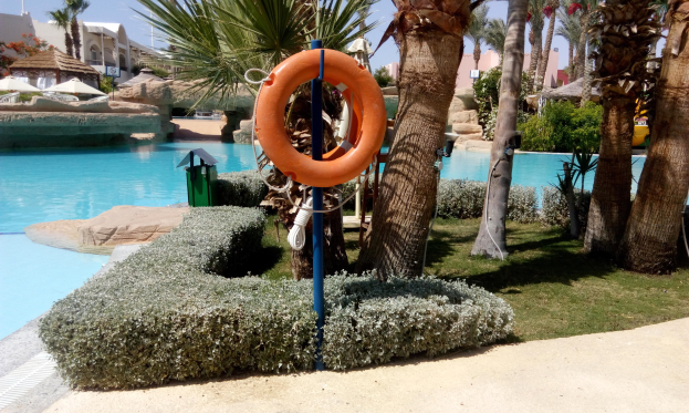 Ein Schwimmbad mit einem Rettungsring in der Mitte, umgeben von Grünflächen und Bäumen, mit Gebäuden und Sonnenschirmen im Hintergrund unter einem klaren blauen Himmel.
