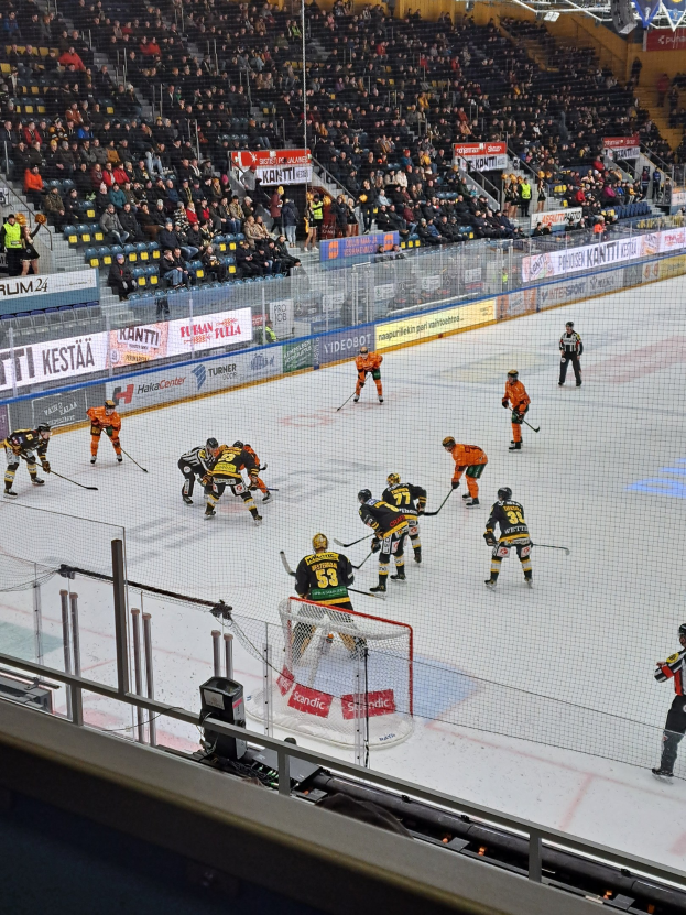 Eine Gruppe von Menschen, die Hockey auf einer Indoor-Eisfläche spielen, umgeben von einem Zaun, mit Zuschauern im Hintergrund und beleuchteter Stadionbeleuchtung oben.
