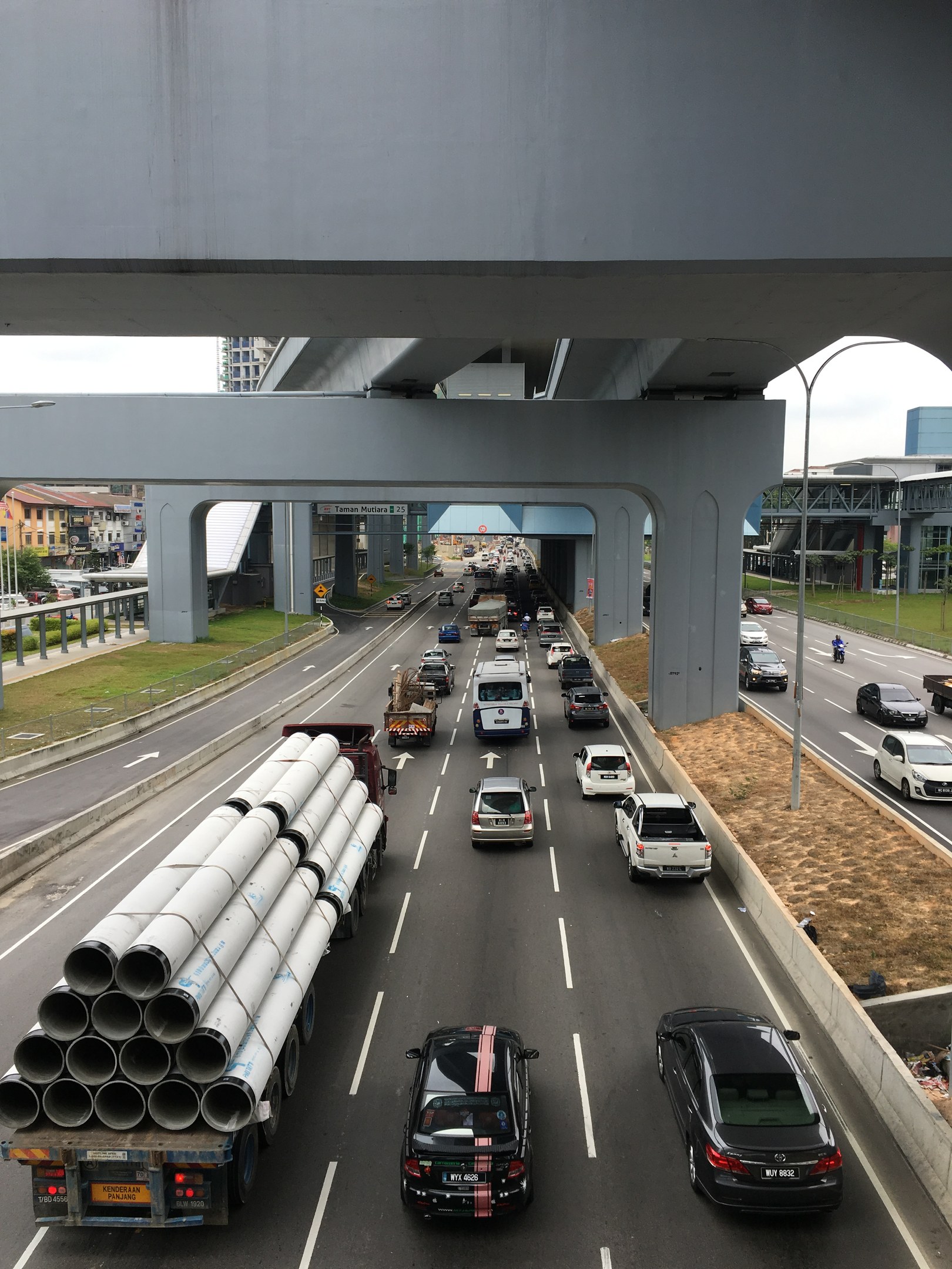 Eine belebte Autobahn mit zahlreichen Fahrzeugen, Straßenlaternen und Infrastruktur unter einer Brücke, mit Gebäuden, Bäumen und Gras im Hintergrund unter einem klaren Himmel.