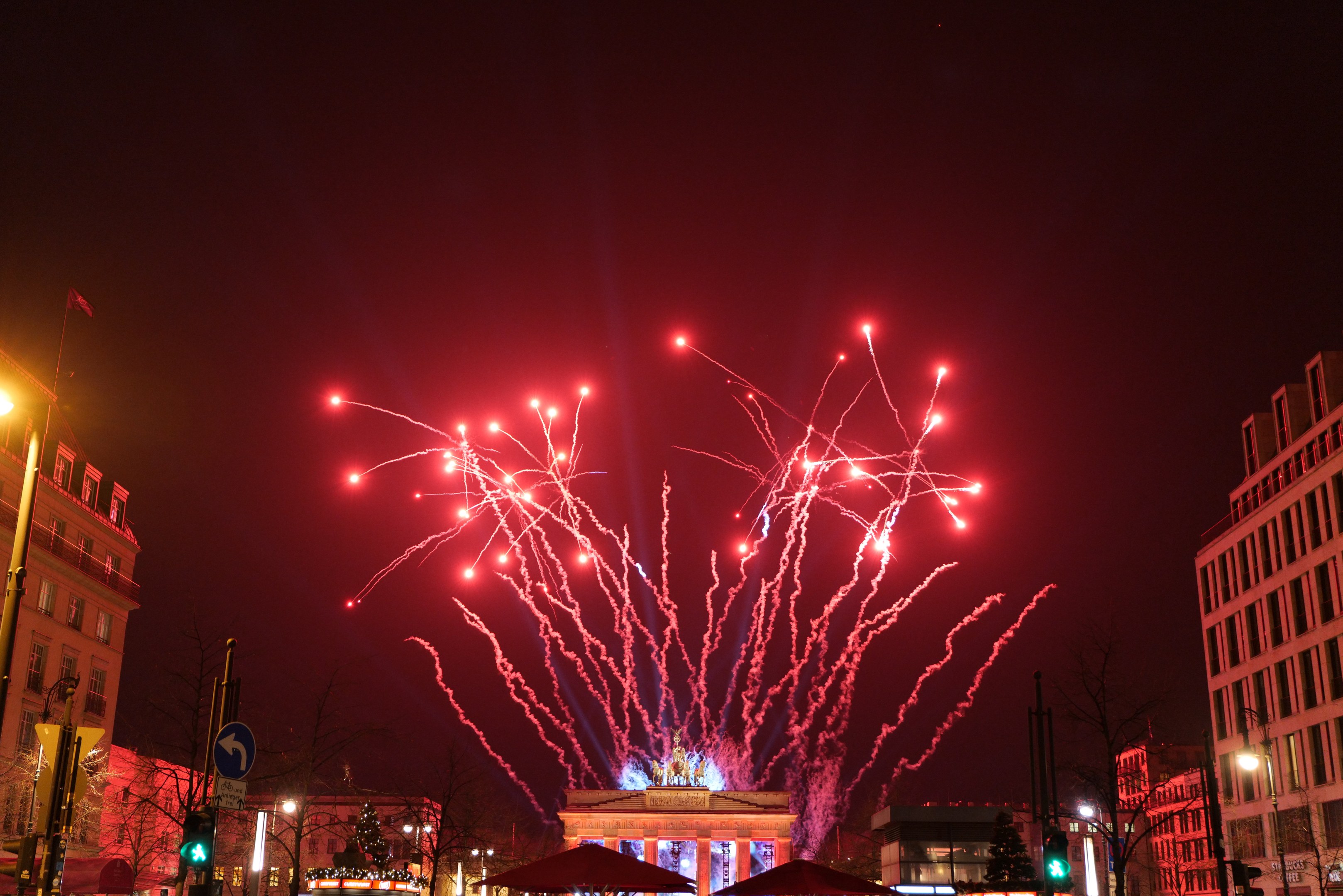 Eine belebte Stadtstraße an einem Silvesterabend in Berlin mit Gebäuden, Bäumen, Laternen, Verkehrszeichen, Schildern, Zelten, Menschen und einem prächtigen Feuerwerk am Himmel.