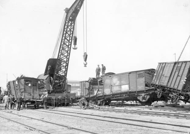 Schwarze und Weiße Szene einer Zugkatastrophe mit einem Kran, der ein Waggon auf die Schienen hebt, mehrere Menschen um den Kran herumstehend, Gebäude und ein klarer Himmel im Hintergrund.