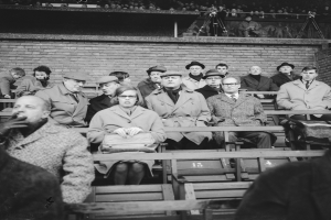 Eine Gruppe von Menschen, sowohl Männer als auch Frauen, sitzt in einem Stadion mit einer Wand und einigen stehenden Personen im Hintergrund, dargestellt in Schwarz-Weiß.