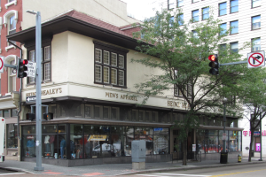 Eine belebte Straßenecke mit einem geparkten Bus, Gebäuden mit Fenstern, Bäumen, Laternen, Verkehrsampeln, Schildern und einem Kaufhaus mit durch die Fenster sichtbaren Schaufensterpuppen.