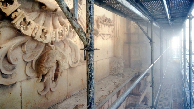 Ein renoviertes Gebäude mit detaillierten Schnitzereien an der linken Wand, Metallstangen an der rechten und einem Gerüst drumherum.