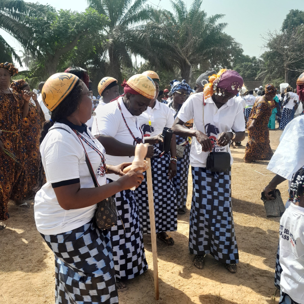 Gruppe von Frauen in weißen und schwarzen Kleidern mit einigen, die gelbe Helme tragen, die auf einem Feld in Ghana stehen, mit Bäumen und einem klaren blauen Himmel im Hintergrund.