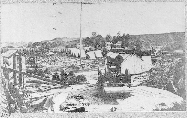 Schwarz-weißes Foto einer Baustelle mit einer Maschine im Vordergrund, umgeben von Hütten, Bäumen, Hügeln und Himmel im Hintergrund, und Text unten lesbar "Die Bauarbeiten an einer neuen Brücke in der Stadt San Francisco, Kalifornien."