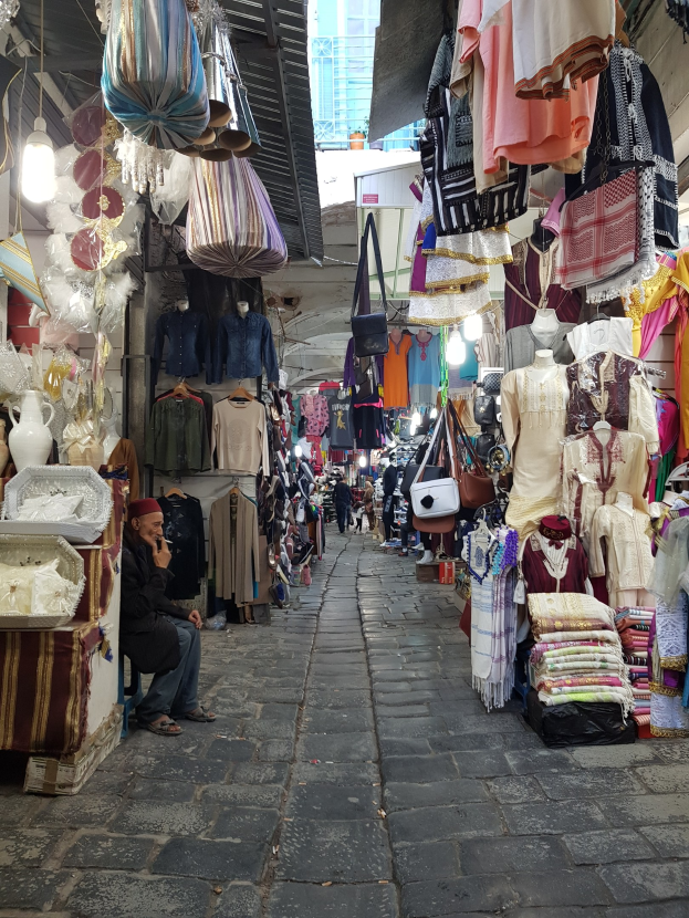 Ein belebter Straßenmarkt in der Altstadt von Jerusalem mit Ständen, die Kleidung, Taschen, Lampen und andere Artikel verkaufen, während Menschen im Hintergrund durch die Gassen schlendern und einkaufen.