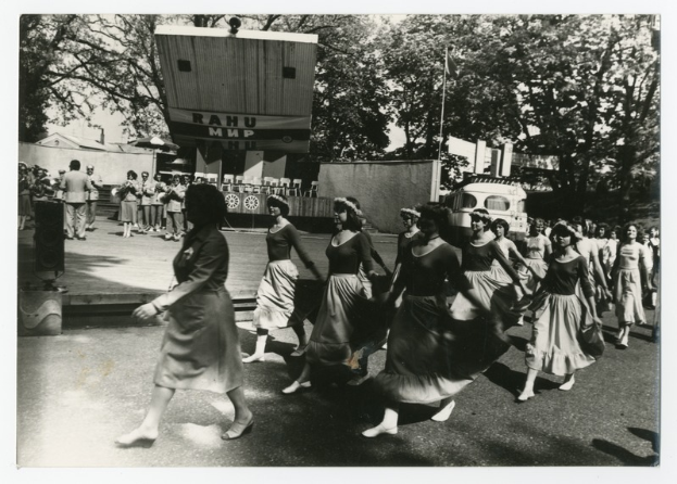 Eine Gruppe von Frauen tanzt auf der Straße, umgeben von einer Menge, einem Lautsprecher, einem Fahrzeug, einer Tafel mit Text und einem Fahnenmast.