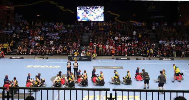 Eine Gruppe von Rollstuhlfahrern bei den Invictus Games Düsseldorf 2020, mit einer großen Zuschauermenge im Hintergrund, einer Treppe, die zu einem Bildschirm führt, und Informationsschildern rund um den Platz.