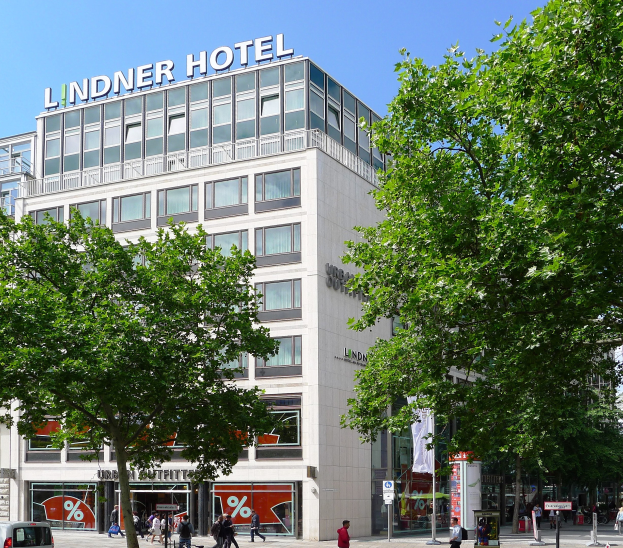 Großes modernes Hotelgebäude mit Glasfenstern, der Name "Lindner Hotel" oben angezeigt, umgeben von Bäumen, Fahrzeugen, Fußgängern und einem sichtbaren Himmel.