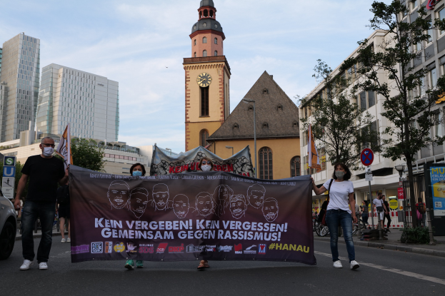 Eine Gruppe von Menschen mit Masken, die eine Straße entlanggehen und eine Fahne mit Text und Bildern halten, mit einem geparkten Auto auf der linken Seite, Gebäuden, Bäumen, Schildern, Polen, einem Uhrenturm und einem klaren blauen Himmel im Hintergrund.