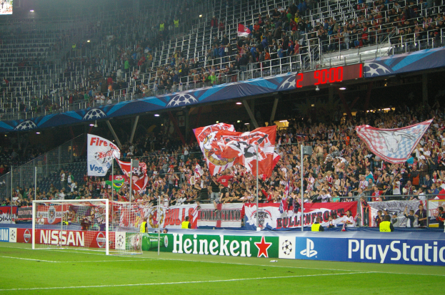 Ein Fußballspiel wird in einem hell erleuchteten Stadion mit einer großen Zuschauermenge, einem Tor, Bannern und einem Bildschirm in der linken oberen Ecke gespielt.
