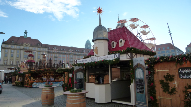 Ein geschäftiger Weihnachtsmarkt auf einem Stadtplatz mit geschmückten Bäumen, Gebäuden, Fahrzeugen, Passanten, Straßenlaternen und einer bewölkten Himmel.