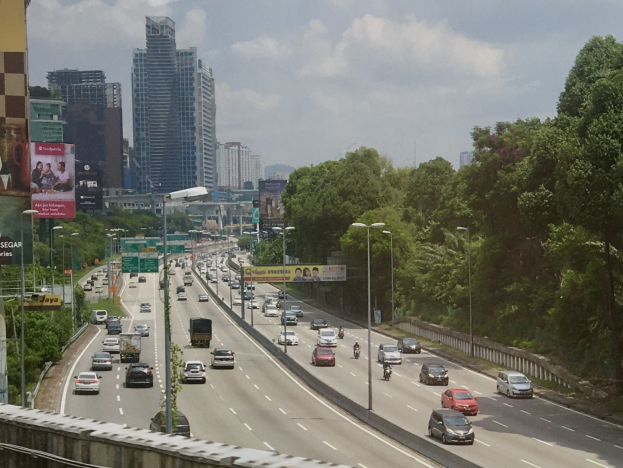 Eine belebte Autobahn mit hohen Gebäuden, Straßenlaternen, Schildern, Bäumen und wolkenverhangenem Himmel.