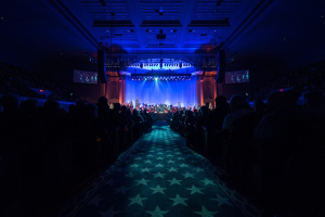Ein großer Zuschauerraum mit Sitzplätzen, eine Bühne mit Musikern und Deckenleuchten und -fernseher, was auf ein Konzerthalle während der Nationalkonvention 2019 hindeutet.